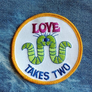 May include: A white embroidered patch with a yellow border. The patch features two green worms with a single blue eye, intertwined with the text "LOVE TAKES TWO" in pink and blue lettering.