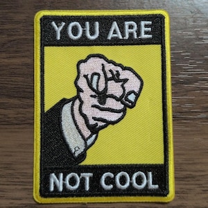 May include: A rectangular embroidered patch with a yellow background and black borders. The patch features a graphic of a hand pointing forward, wearing a dark suit sleeve. The text YOU ARE is at the top and NOT COOL is at the bottom in white.