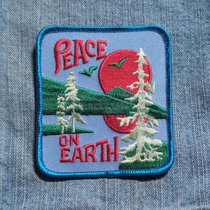May include: A square embroidered patch with a light blue background featuring a red sun, green hills, and white pine trees. The text PEACE ON EARTH is stitched in red lettering. The patch has a dark blue border and rounded corners.