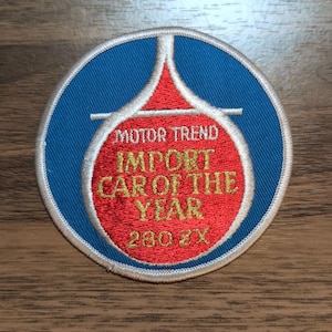 May include: A circular embroidered patch featuring a blue background with a central red teardrop shape outlined in white. The text MOTOR TREND IMPORT CAR OF THE YEAR 280 ZX is stitched in gold thread across the red section of the circular patch.