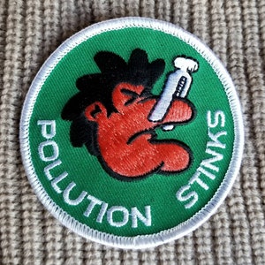 May include: A green and white round patch with a cartoon character holding a cigarette. The text on the patch reads "Pollution Stinks".