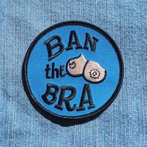 May include: A circular embroidered patch with a bright blue background and a black stitched border. The text BAN the BRA is embroidered in black letters. Two beige, rounded shapes with small floral details are positioned in the center.