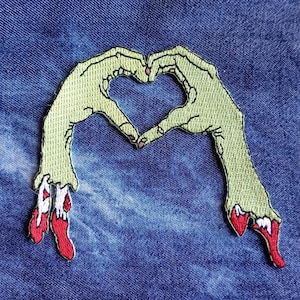 May include: Green embroidered patch of two hands forming a heart shape with red dripping blood from the fingertips.