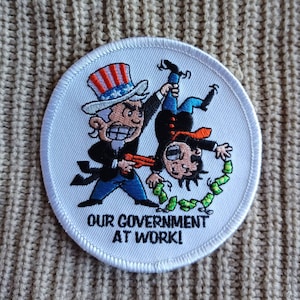 May include: White embroidered patch with a cartoon image of Uncle Sam holding a man upside down by the leg. The man is tied with a string of money. The text "OUR GOVERNMENT AT WORK!" is below the image.