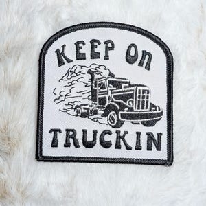 May include: A white embroidered patch with a black border featuring a black graphic of a semi-truck emitting exhaust smoke. The text "KEEP ON" is arched above the truck, and the word "TRUCKIN" is positioned below the truck in black lettering.