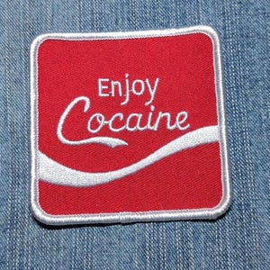 May include: A square embroidered patch with a red background and a white border. The text Enjoy Cocaine is stitched in white across the center, accompanied by a white curved line underneath the word Cocaine, mimicking a classic script style.
