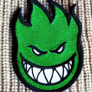 May include: Green and black embroidered patch with a cartoon flame-shaped face with a wide, toothy grin and angry eyebrows.