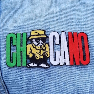 May include: An embroidered patch featuring the word CHICANO in bold block letters. The letters C and H are green, A and N are white, and O is red. A cartoon character wearing a yellow hat, yellow shirt, and black pants is positioned between the letters.