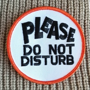 May include: A white round patch with an orange border. The patch has black embroidered text that reads "PLEASE DO NOT DISTURB".