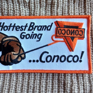 May include: A rectangular embroidered patch with an orange border on a beige knit background. The white patch features a brown gloved hand holding a thin line connected to an orange triangle with the text Conoco and the phrase Hottest Brand Going ...Conoco!