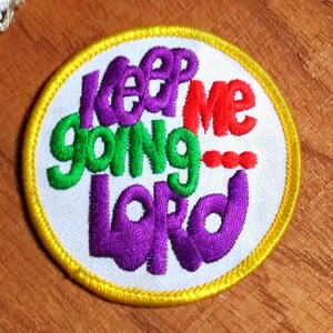 May include: Embroidered patch with the words "Keep Me Going Lord" in purple, green, and red. The patch has a white background, a yellow border, and is circular in shape. The patch is on a wooden surface.