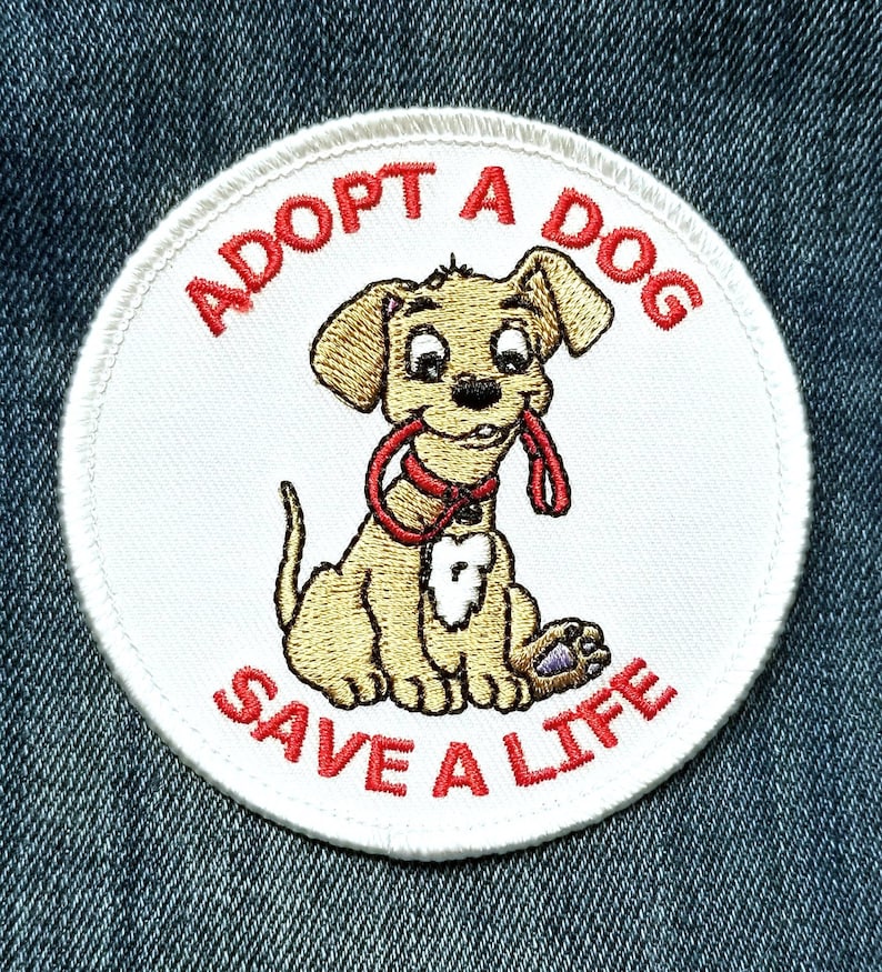 May include: A circular embroidered patch with a white background featuring a tan puppy holding a red leash in its mouth. The text ADOPT A DOG is arched above the puppy, and the text SAVE A LIFE is arched below, both in red capital letters.