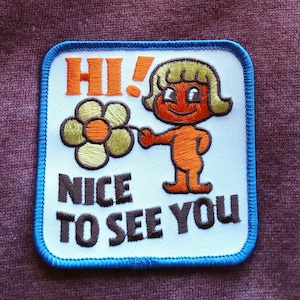 May include: A white embroidered patch with a blue border. It features a cartoon character holding a flower and the text "Hi! Nice to see you."