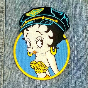 May include: A blue and yellow embroidered patch featuring a cartoon character with a black cap, blue eyes, and a leopard print top.