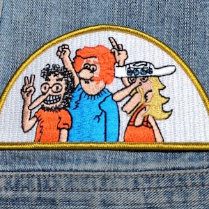 May include: A semi-circular embroidered patch featuring three cartoon characters on a white background with a gold border. The left character has dark hair and glasses, the center has orange hair, and the right wears a white hat with blue accents.