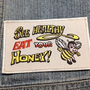 May include: A rectangular embroidered patch with a white background and a stitched border. The text reads "BEE HEALTHY EAT YOUR HONEY!" in yellow and red thread. A cartoon bee with yellow and black stripes and white wings is on the right side.
