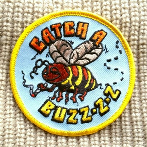 May include: A blue and yellow embroidered patch with a cartoon bee smoking a cigarette. The text "Catch a Buzz-z-z" is embroidered in yellow on the patch.