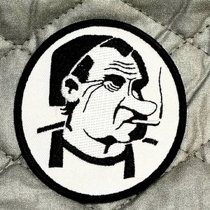 May include: Black and white embroidered patch of a cartoon man with a large nose smoking a cigarette. The man is wearing a collared shirt and has a serious expression.