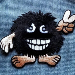 May include: Black fuzzy monster with a big smile and white teeth, wearing brown shoes and giving a peace sign with one hand.