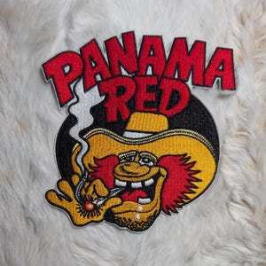 May include: Embroidered patch featuring a cartoon character with a yellow hat and red hair, smoking. The patch has the words "PANAMA RED" in red, outlined in white. The background is black and the patch is on a white, fluffy surface.