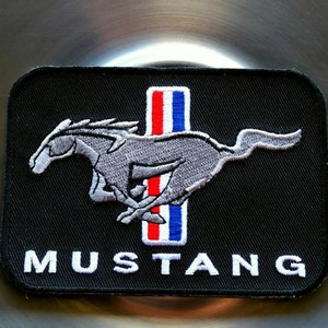 May include: Black rectangular patch featuring the word "MUSTANG" in white, with a running gray horse emblem. A vertical stripe with red, white, and blue colors is on the right side of the horse.