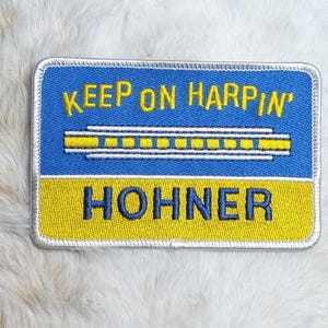 May include: A rectangular embroidered patch featuring a blue top section with the yellow text KEEP ON HARPIN' above a yellow harmonica graphic. The bottom section is yellow with the blue text HOHNER. The patch has a silver-colored border.