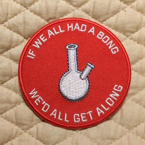 May include: A circular red embroidered patch featuring a white stitched graphic of a water pipe in the center. The text "IF WE ALL HAD A BONG WE'D ALL GET ALONG" is embroidered in white capital letters around the perimeter of the red circular patch.