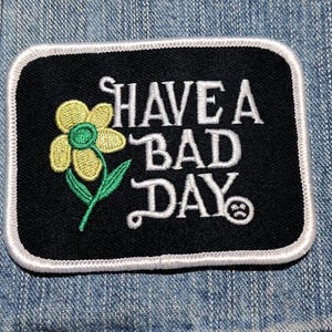 May include: A rectangular embroidered patch with a black background and a white border. It features a yellow flower with a green stem and the white embroidered text HAVE A BAD DAY with a small sad face icon at the end of the final word.