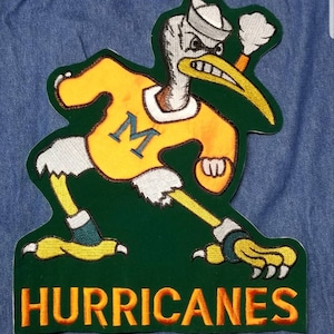 May include: A green and yellow embroidered patch featuring a cartoon bird wearing a sailor hat and holding a football. The bird is wearing a yellow jersey with a large "M" on the front. The text "HURRICANES" is embroidered in orange below the bird.