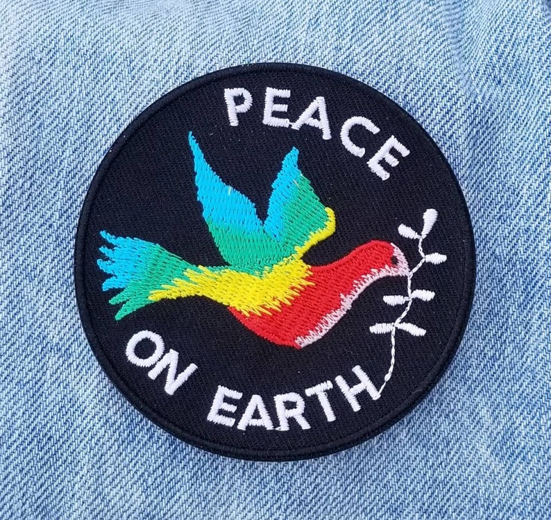 May include: A black and white embroidered patch with a colorful dove carrying an olive branch. The patch reads "Peace on Earth".