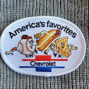 May include: White oval embroidered patch with the text "America's Favorites" in black. The patch features a cartoon baseball, hot dog, and slice of pie with the text "Chevrolet" in red and blue below.