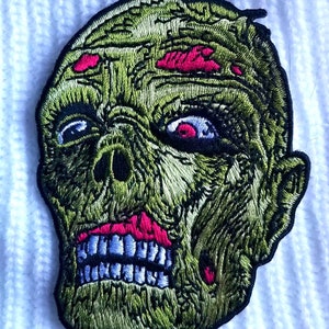 May include: Embroidered patch of a green zombie face with pink and blue details. The zombie has a large, open mouth with visible teeth.