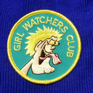May include: A yellow and teal embroidered patch with the text "Girl Watchers Club" and a cartoon image of a hand with a tongue sticking out.