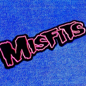 May include: A black patch with the word "MISFITS" in a stylized font, outlined in pink. The patch is set against a blue denim background, creating a contrast between the dark text and the bright fabric.