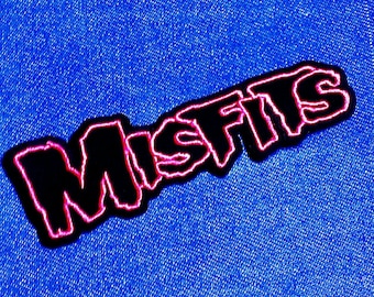 May include: A black patch with the word "MISFITS" in a stylized font, outlined in pink. The patch is set against a blue denim background, creating a contrast between the dark text and the bright fabric.