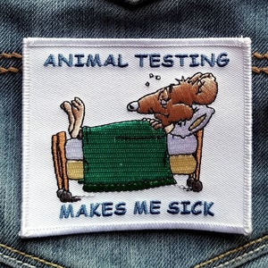 May include: A square embroidered patch with a white background featuring a brown mouse lying in a bed with a green blanket. The text ANIMAL TESTING is embroidered in blue at the top, and the text MAKES ME SICK is embroidered in blue at the bottom.