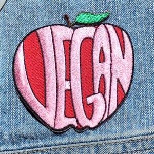 May include: An embroidered patch shaped like an apple with a red background and light pink letters spelling out the word VEGAN. The apple features a small green leaf at the top and is outlined in black thread, attached to a blue denim fabric.