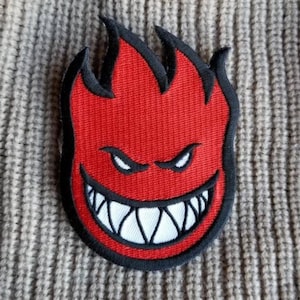 May include: An embroidered patch featuring a stylized red flame character with a black outline. The character has narrow, aggressive white eyes and a wide, toothy grin with sharp white teeth. The patch is placed on a textured, ribbed beige fabric.