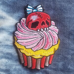 May include: A red skull with a pink and yellow cupcake frosting and a blue and white bow on top. The cupcake is embroidered on a denim fabric.
