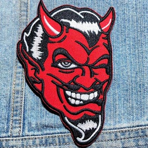 May include: An embroidered patch featuring a stylized red devil face with black hair, horns, and a goatee. The devil has a wide, toothy grin and one eye is winking. The design is outlined in black thread, creating a bold, graphic appearance.