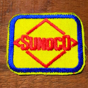 May include: A rectangular embroidered patch with rounded corners featuring a bright yellow background, a thick blue border, and a red diamond shape in the center. The word SUNOCO is embroidered in red capital letters across the center diamond.