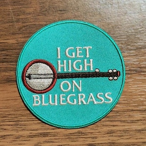 May include: A circular embroidered patch with a teal background featuring a white banjo graphic with a red rim. The text I GET HIGH ON BLUEGRASS is stitched in white across the center, positioned above and below the banjo neck illustration.