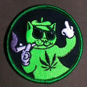 May include: Green embroidered patch of a cartoon cat wearing sunglasses, smoking a pipe, and giving the middle finger. The cat is wearing a green shirt with a marijuana leaf on it.