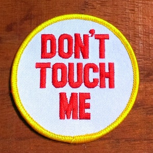 May include: A round patch with the words "DON'T TOUCH ME" embroidered in red on a white background. The patch has a yellow border and is likely designed to be sewn onto clothing or accessories.