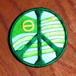 May include: Embroidered peace sign patch featuring a green outline and a green peace symbol. The interior design includes light blue and green horizontal stripes, with a yellow circle and horizontal line. The patch is circular and has a stitched border.