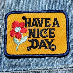 May include: A rectangular embroidered patch with a mustard yellow background and a dark blue border. The patch features a red flower with a light blue center and green stem, alongside the black text "HAVE A NICE DAY" with a small smiley face.
