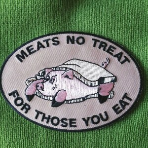 May include: An oval-shaped embroidered patch on a green knit fabric background. The patch features a light tan center with a black border and the black text MEATS NO TREAT FOR THOSE YOU EAT. A pink pig illustration is centered on the patch.