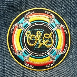May include: A circular embroidered patch featuring a black background with a central yellow cursive script logo. The design includes concentric rings of red, orange, yellow, and light blue, accented by silver metallic thread and a blue geometric pattern.