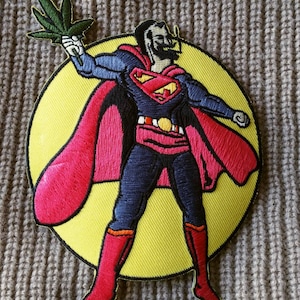 May include: An embroidered patch featuring a superhero holding a marijuana leaf. The superhero is wearing a red cape, blue suit, and yellow belt with the letter 'S' on it. The background is a yellow circle.