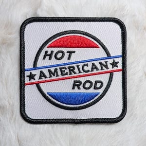 May include: A square embroidered patch with rounded corners and a black border. The design features a white background with a central circle containing red and blue horizontal stripes. The text HOT AMERICAN ROD is embroidered in black.